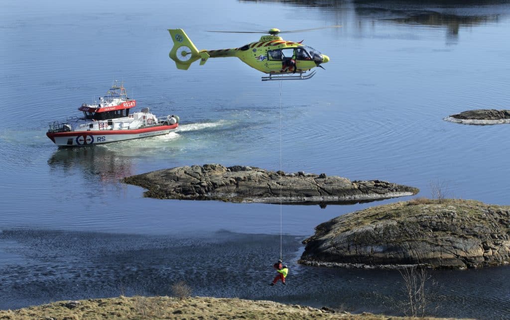 Luftambulansehelikopter med redningsmann og pasient hengende under i tau under oppdrag. Redningsskøyta i bakgrunnen. 