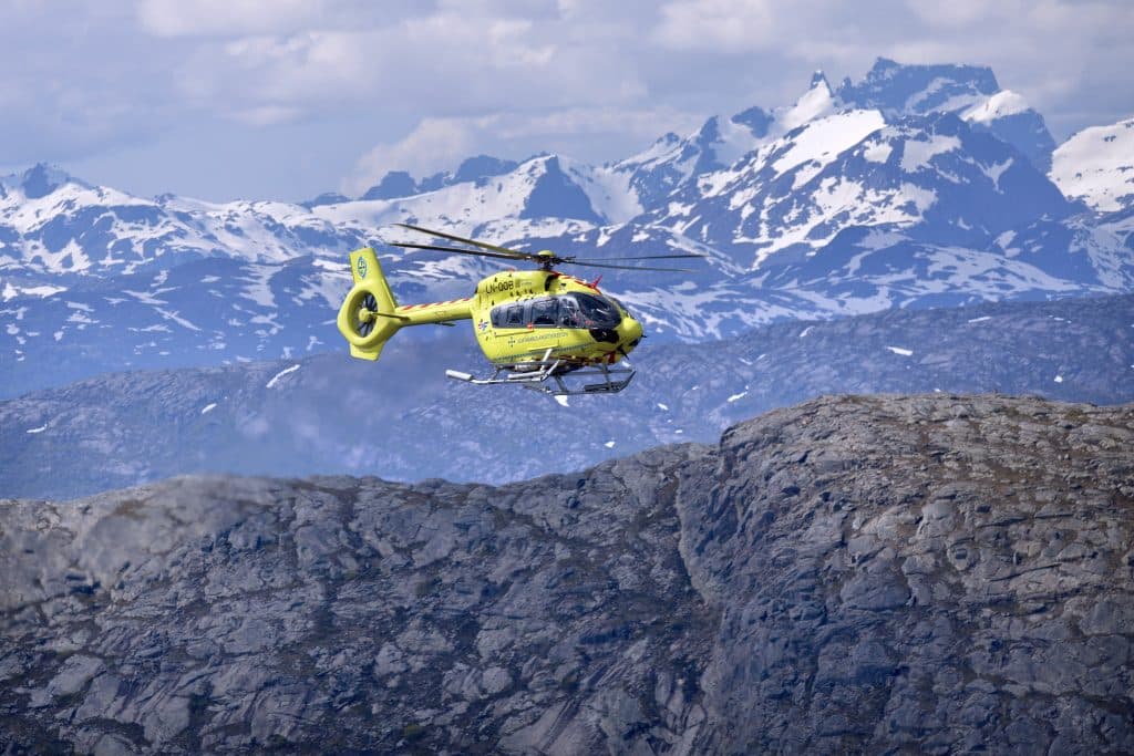 Luftambulansehelikopter flyr over fjellandskap med snødekte topper. 